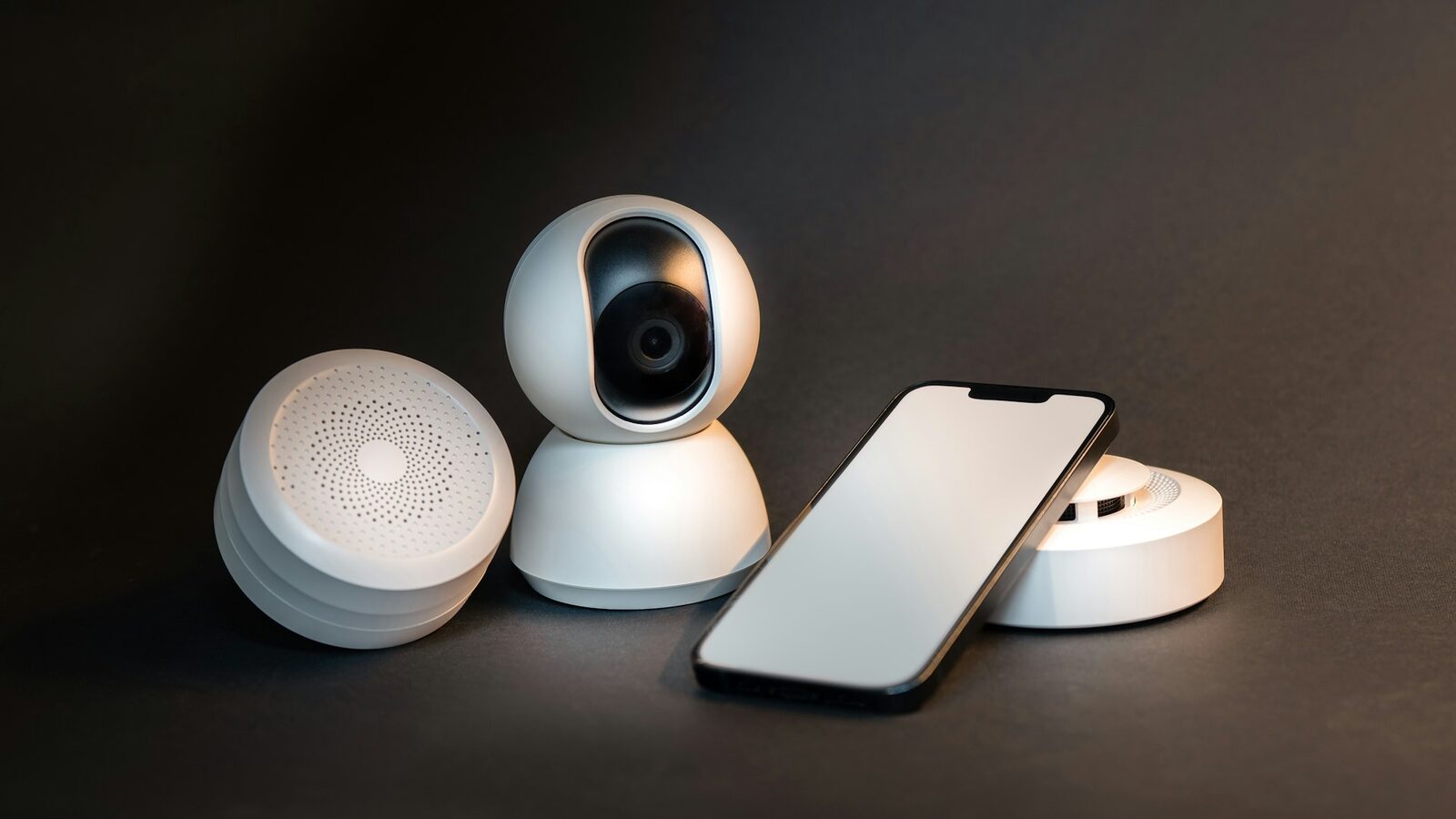 White smart home devices on a dark background: a pan-and-tilt security camera, a smartphone with a blank screen, a hub and a smoke sensor.